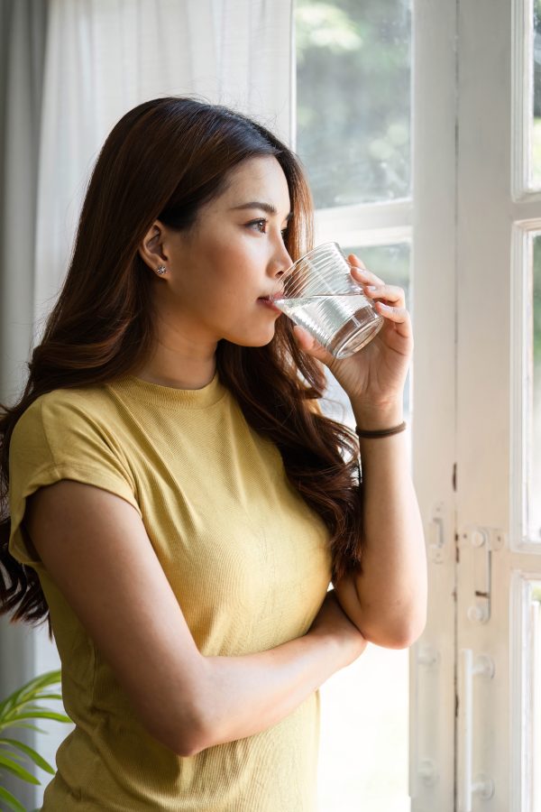 Hydration and Wellness. A woman sips water while contemplating her health by a sunlit window. Water Softener