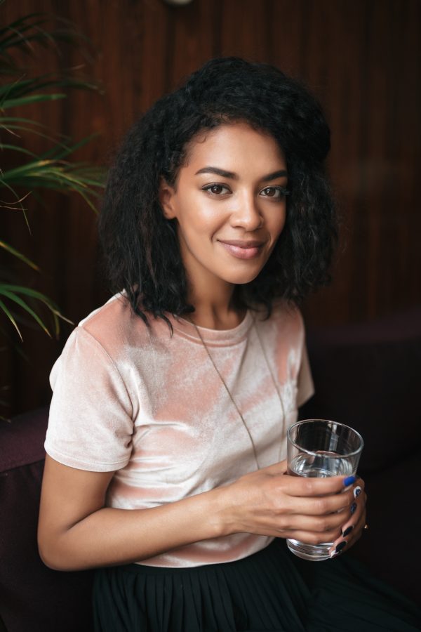 Young beautiful African American girl sitting in restaurant and looking in camera. Smiling lady with dark curly hair sitting in cafe with glass of water in hand Water Softener